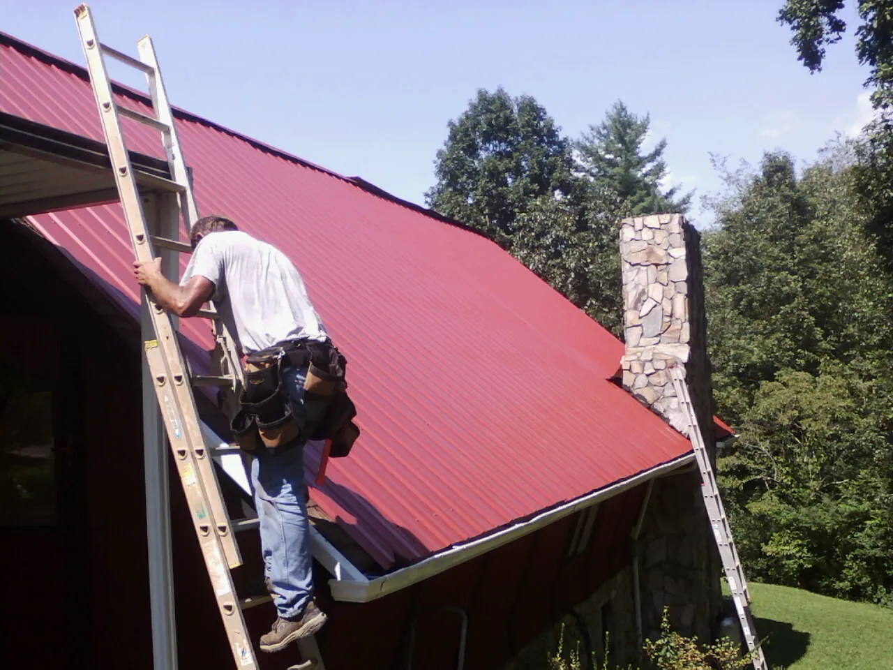 Roofer on a ladder beside a red metal roof and stone chimney — Soffit & Fascia Repair by Roofing Brothers in Boaz